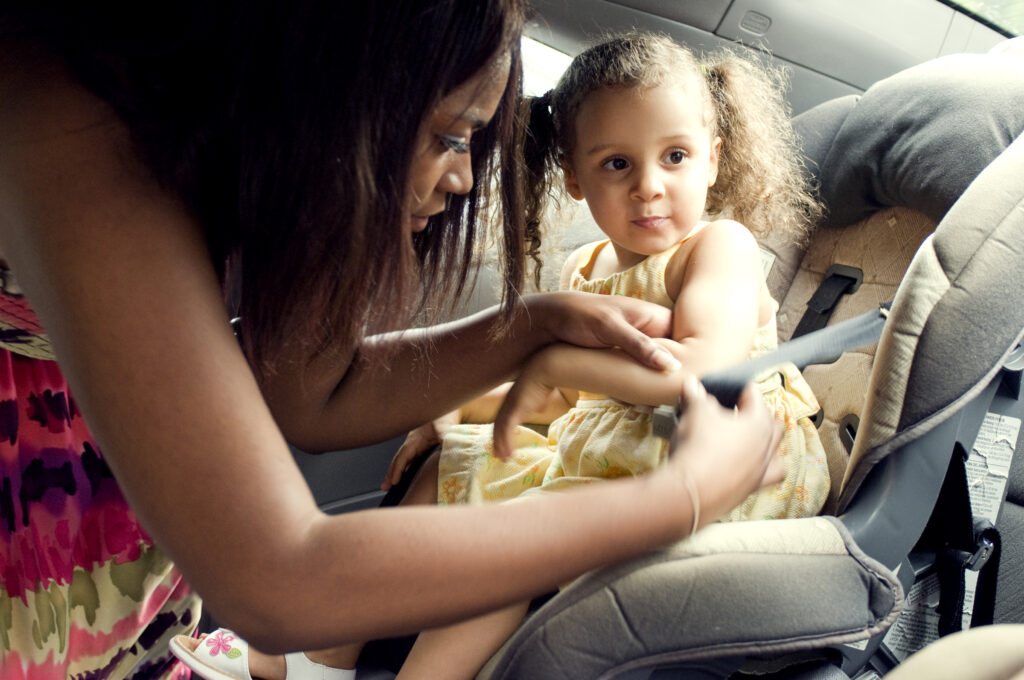 This image depicts a mother in the process of securing her young daughter into her child safety seat, which was located in the vehicle’s rear seat.