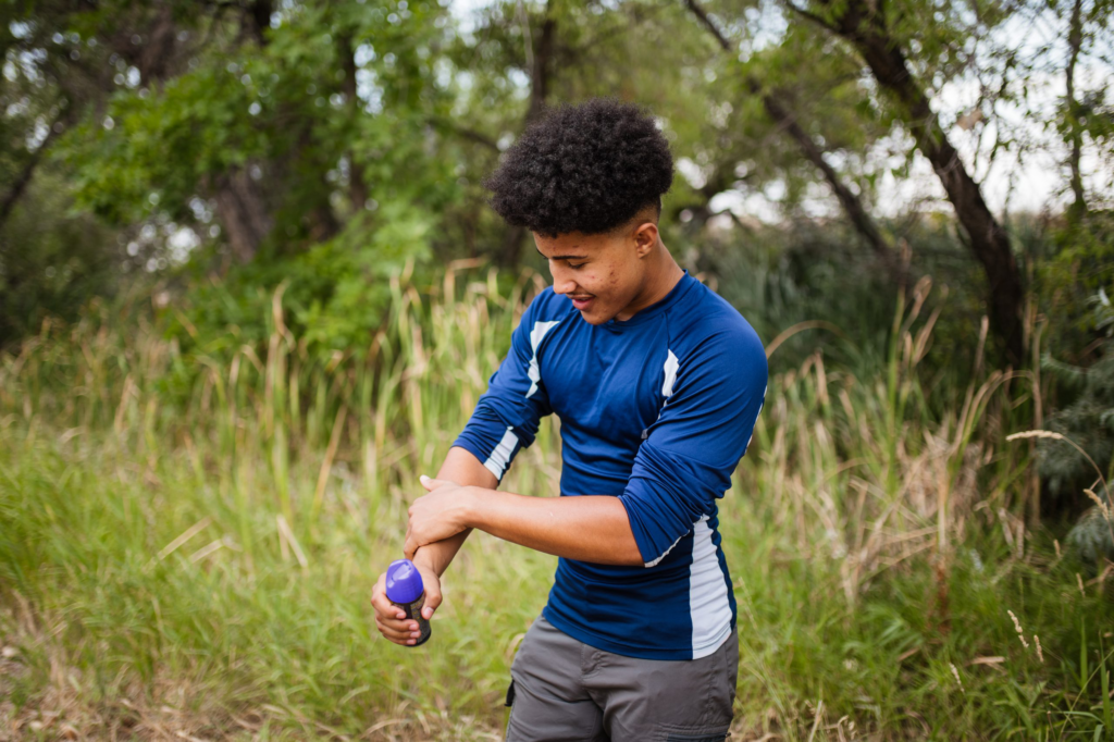 A young Black teen is rubbing insect repellent on his arm. He is outside with high grass behind him.