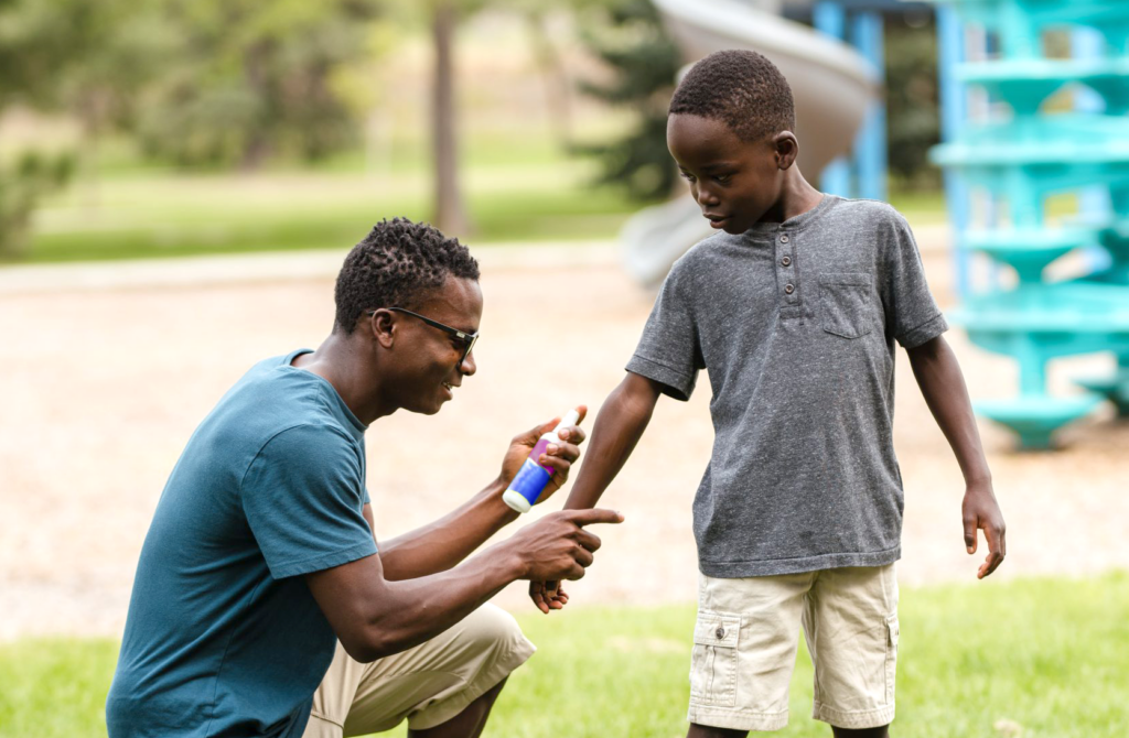 A Black father rubbing bug repellant on his kid's arm. They are in a play ground.