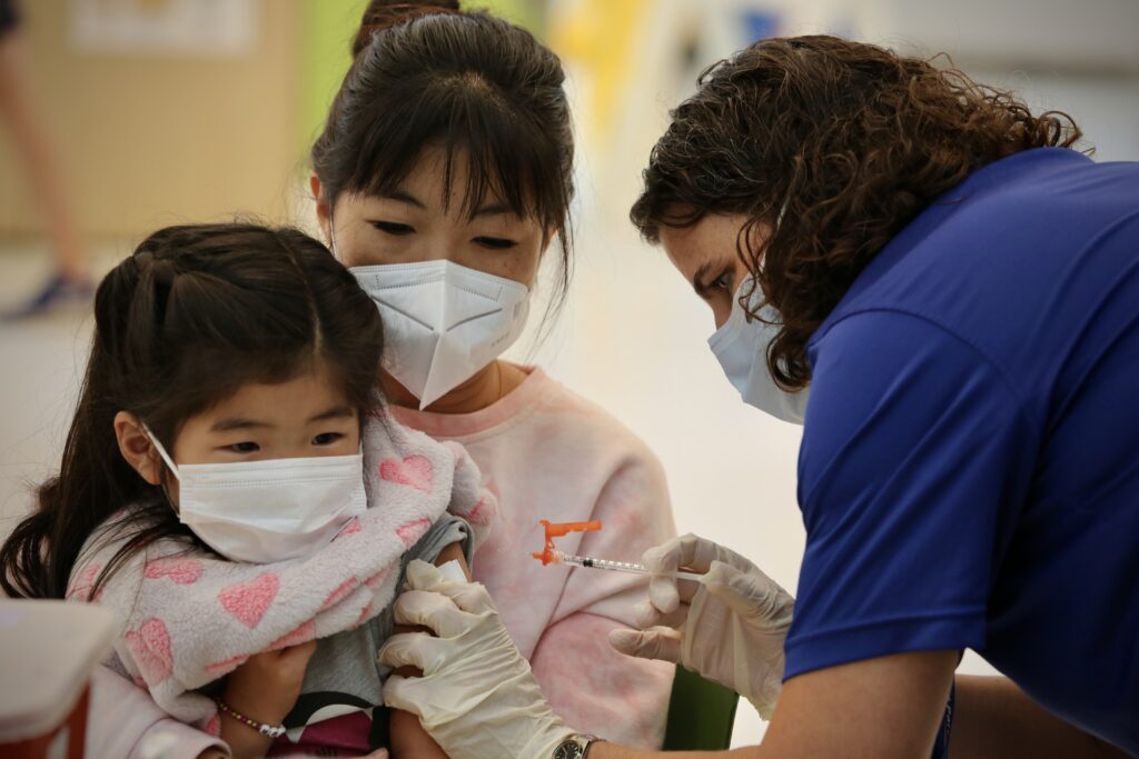 An Asian girl is sitting on her mom's lap. A school nurse who is white is about to administer a vaccine in her left arm.