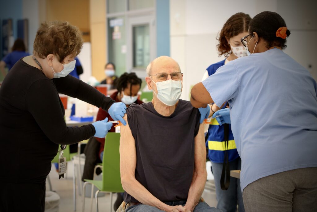 A white elderly man receiving both flu and COVID vaccines from our nurses at a CPHD clinic in fall 2023. Everyone in the photo is wearing a mask.