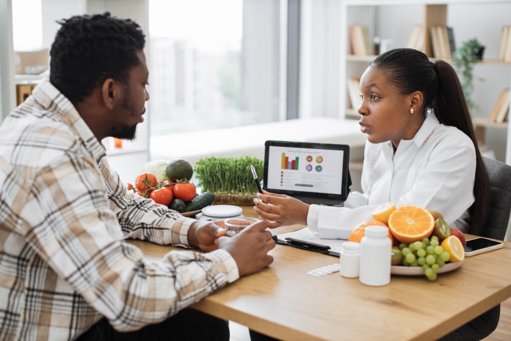 A Black woman professional displaying nutrition data graphically via tablet for a Black man. This is in doctor's workplace. She is sitting next to a bowl of fruit. 