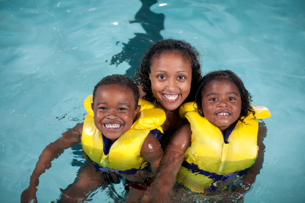 Photo of two Black kids with their mother in the middle. They are all in a pool. The kids have life jackets on. Photo from iStock.