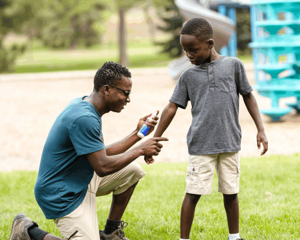 Photo of Black father holding his child’s wrist. He is ready to apply bug spray to his child’s arm. They are outside, it is sunny, and a playground is in the background.
