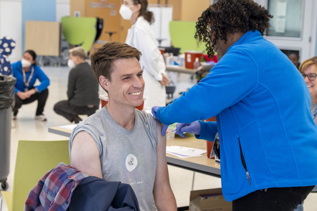 Photo of a white man smiling as he is getting a vaccine in his left arm. He has his gray t-shirt sleeves rolled up to get the vaccine. The vaccine is being given by a School Health Nurse who is Black. She is wearing a blue jacket. There are staff in the background. 