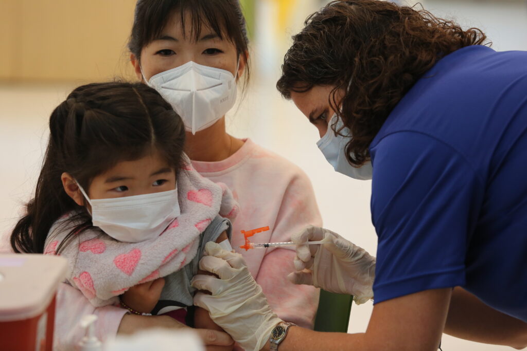 Photo of a young Asian girl being held by her mom. The young girl is receiving a vaccine in her left arm. The vaccine is given by a school nurse. From one of CPHD's vaccine clinic in 2023.