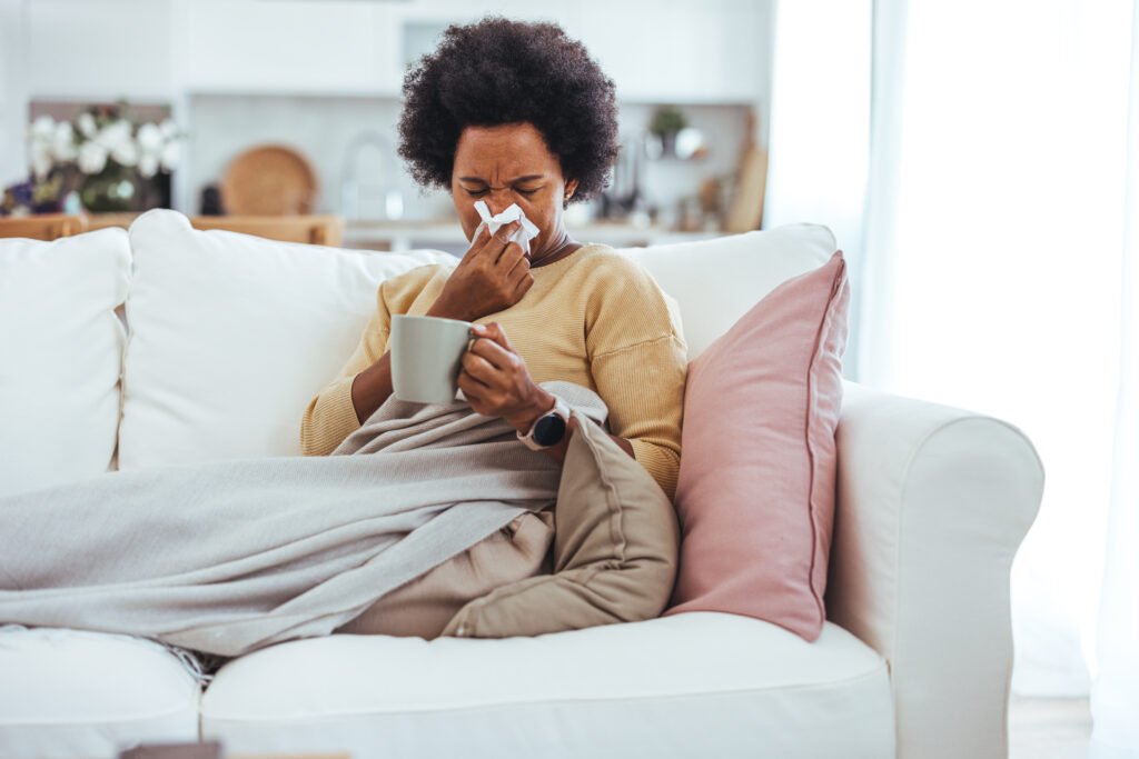 Stock photo of a Black middle aged woman who is blowing her nose. She is holding tea, covering herself with a blanket, and sitting on the couch at home. 