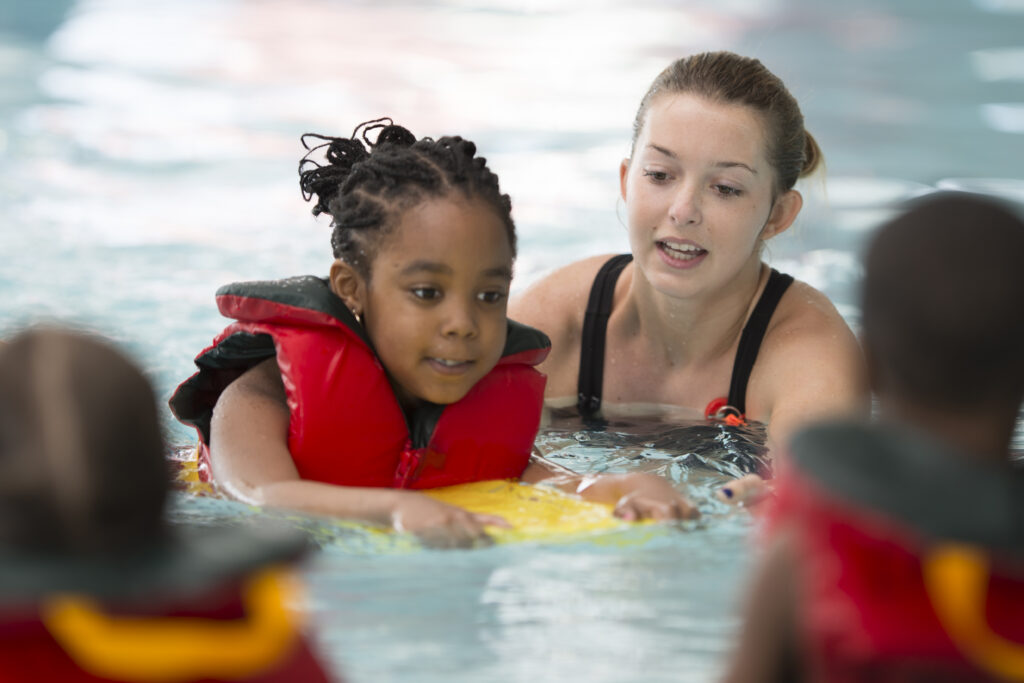 A young Black girl is holding on to a kick board with her swim instructor guiding her. There are two young kids in the pool with them.  