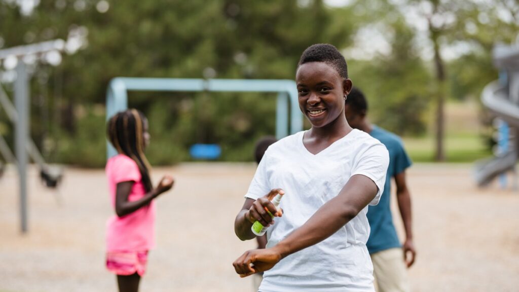 A Black girl is smiling for the camera as she applies bug spray to her arm. She is on a playground with her family.