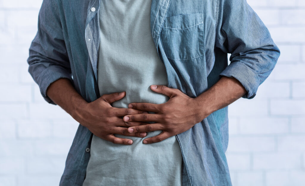 A Black Man Touching His Abdomen Having Painful Stomachache. He's Standing Over Gray Studio Background. Pho