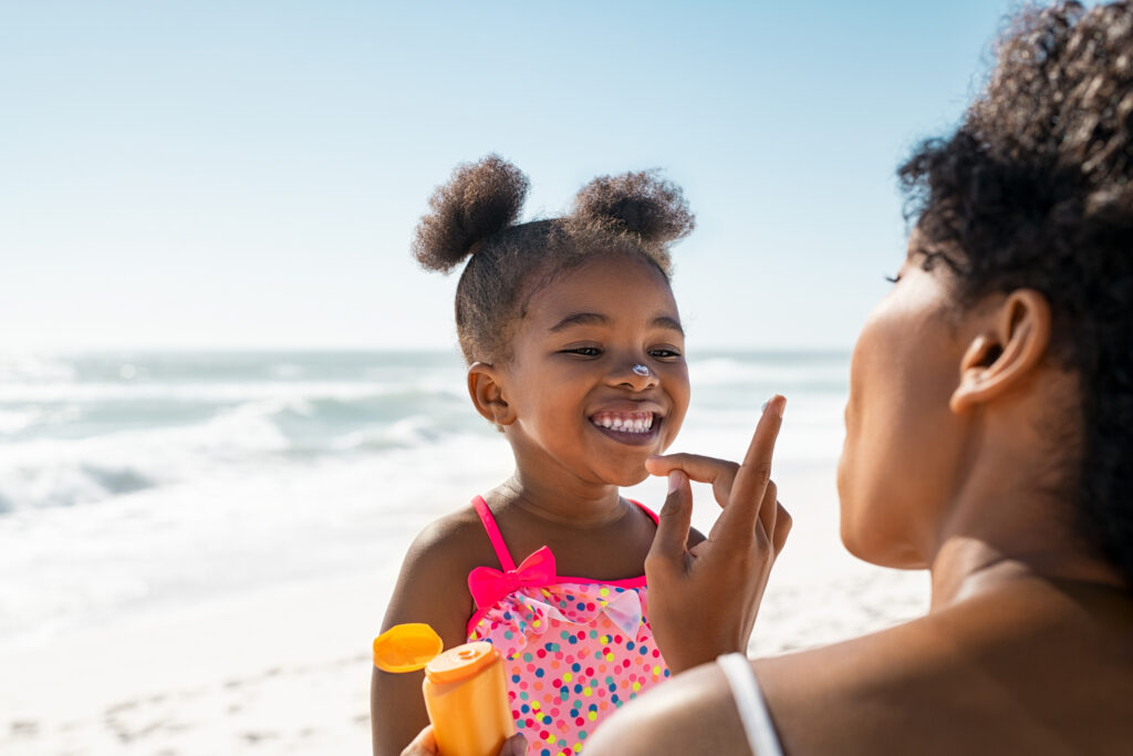 A Black mom putting sunscreen on her young daughter's nose. The daughter is smiling. They are at the beach.