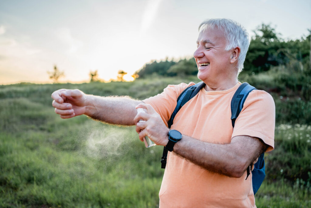 A cheerful White elder man spraying bug spray on his arm. He's in outside in nature. 