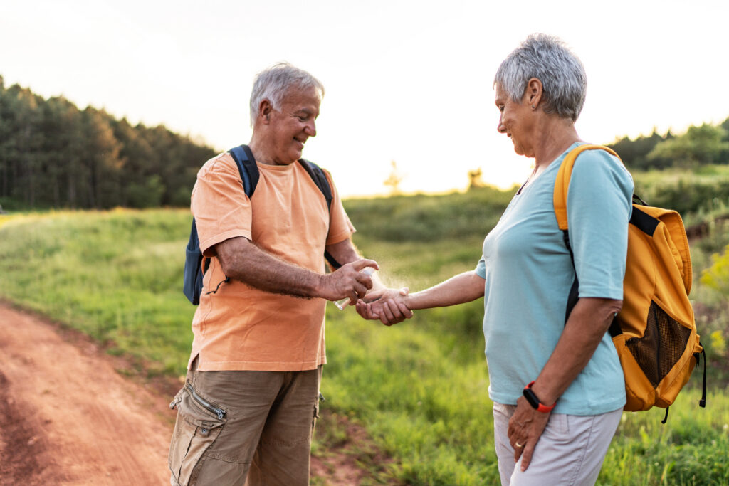 Cheerful senior white man and his wife using mosquito spray in nature. Spraying insect repellent on skin outdoor. 